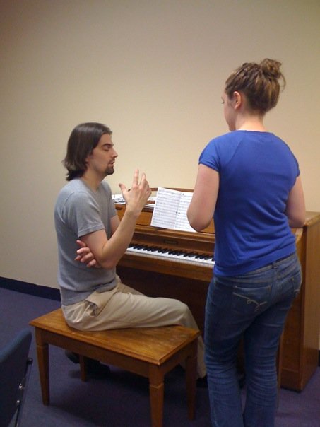 Long haired man sitting at a piano instructing; coaching a female student